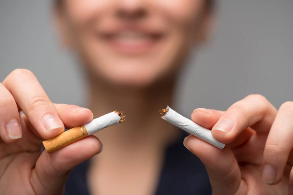 Portrét mladé atraktivní ženy, která rozbíjí cigaretu na kousky. Studiový záběr se selektivním zaostřením izolovaný na šedé ploše